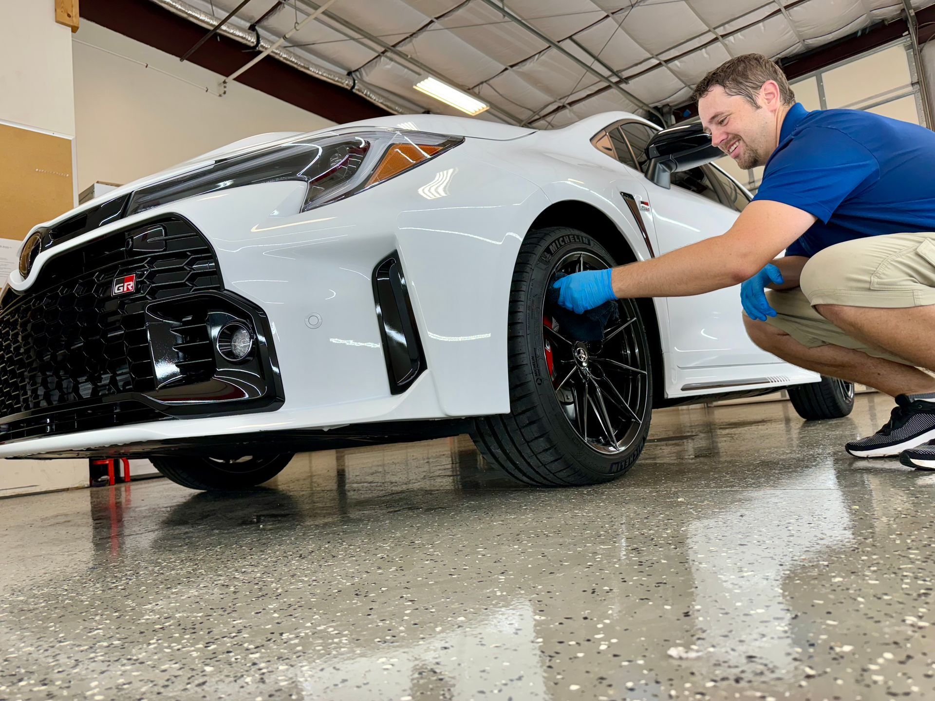 A man is kneeling down next to a white sports car in a garage.