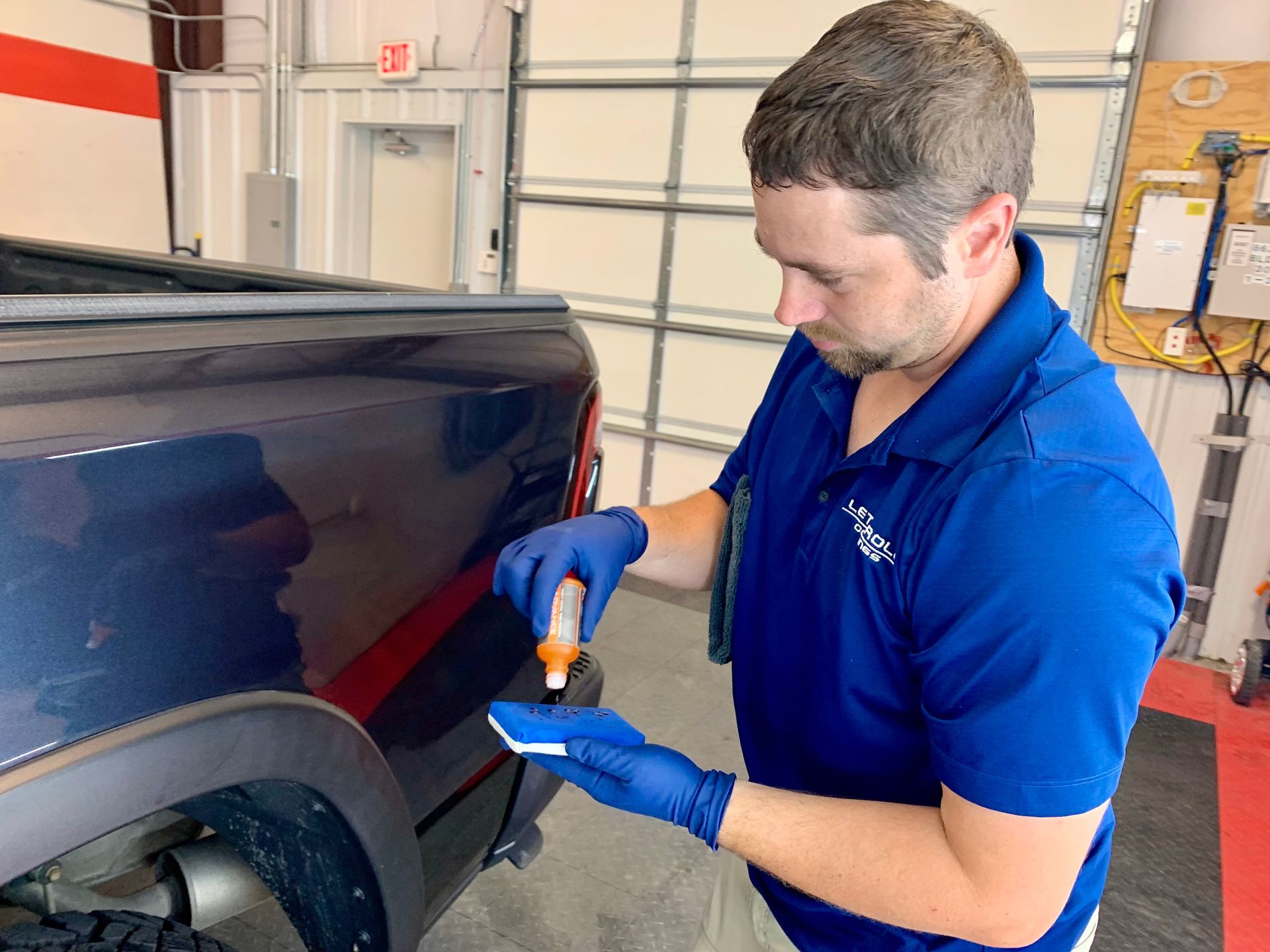 Man in blue shirt and gloves applying product to a blue car panel in a garage.