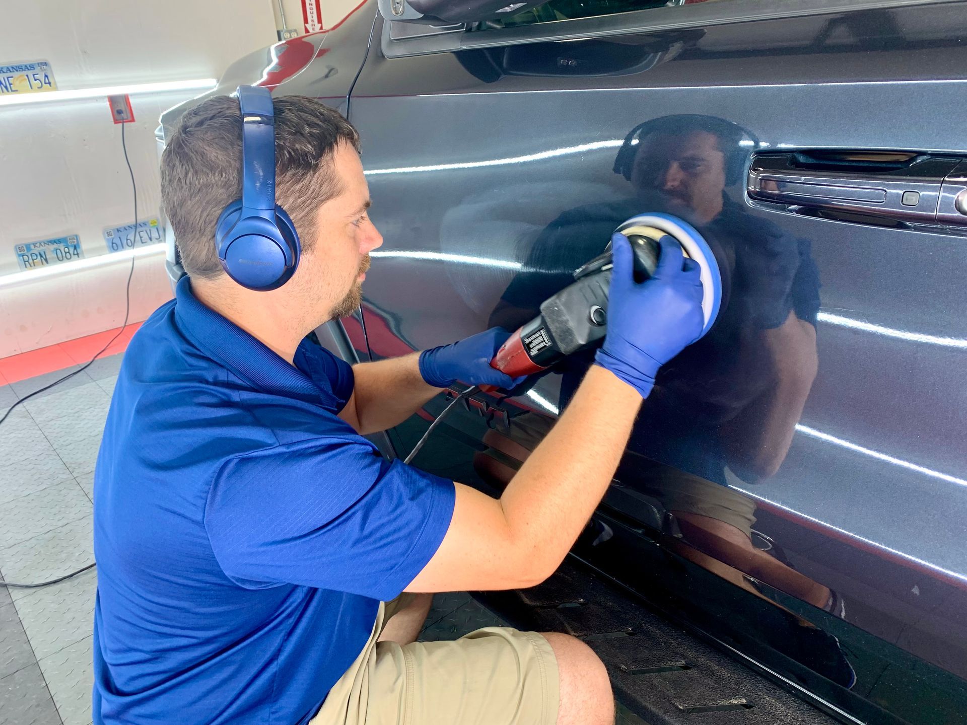 Man polishing a dark car, wearing blue headphones, gloves, and a blue shirt.