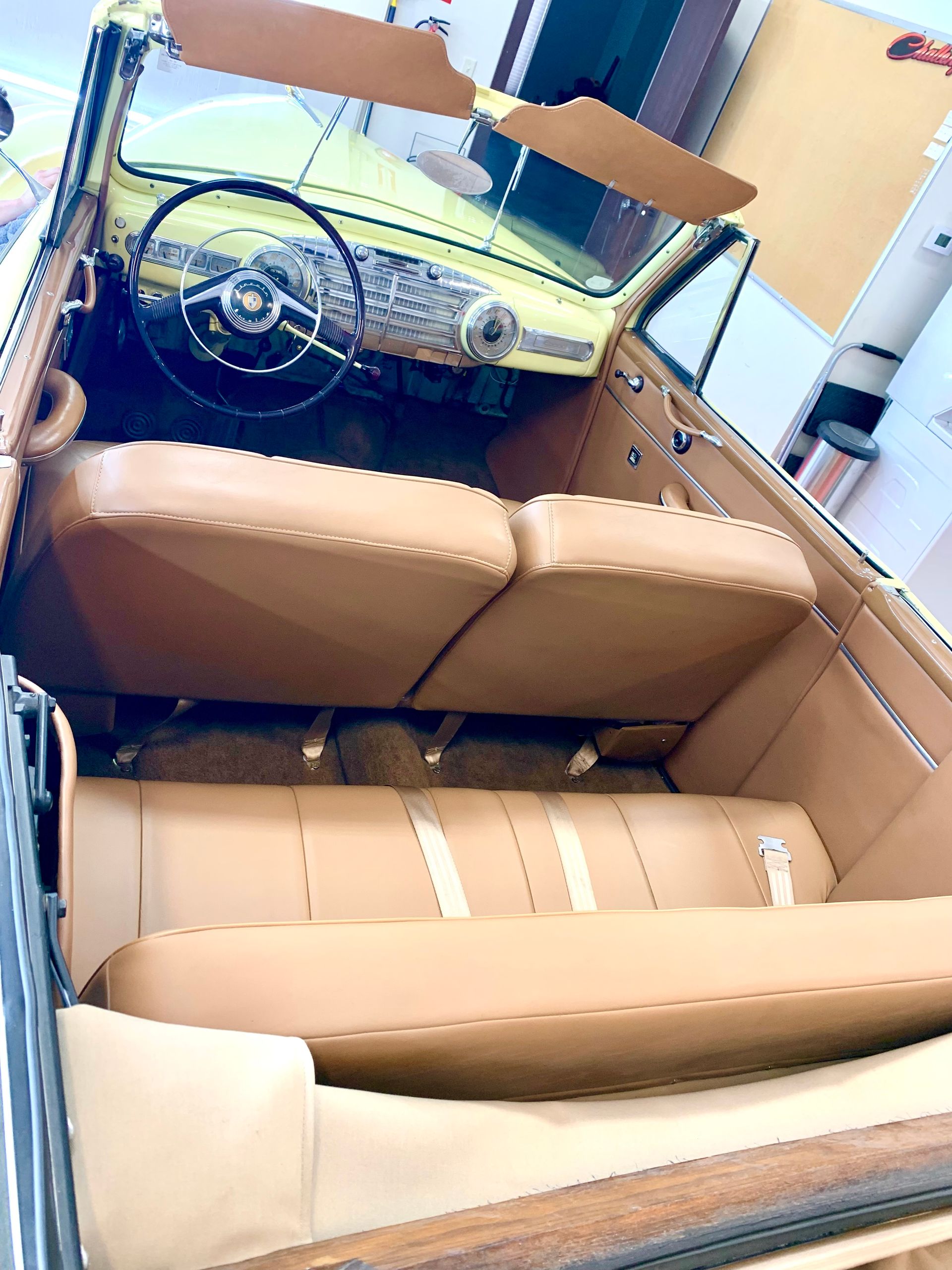 Tan interior of a vintage convertible car, with tan seats, a black steering wheel, and a yellow dashboard.