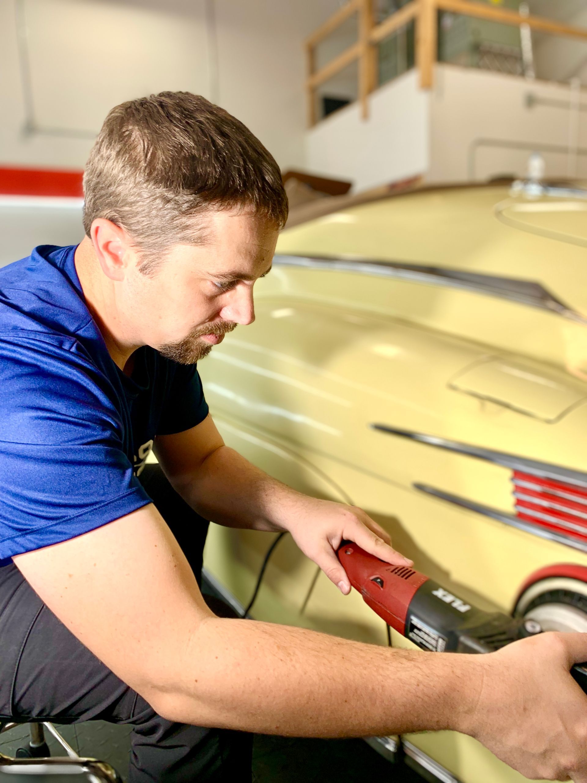 Man polishing a yellow classic car with a power tool in a garage.