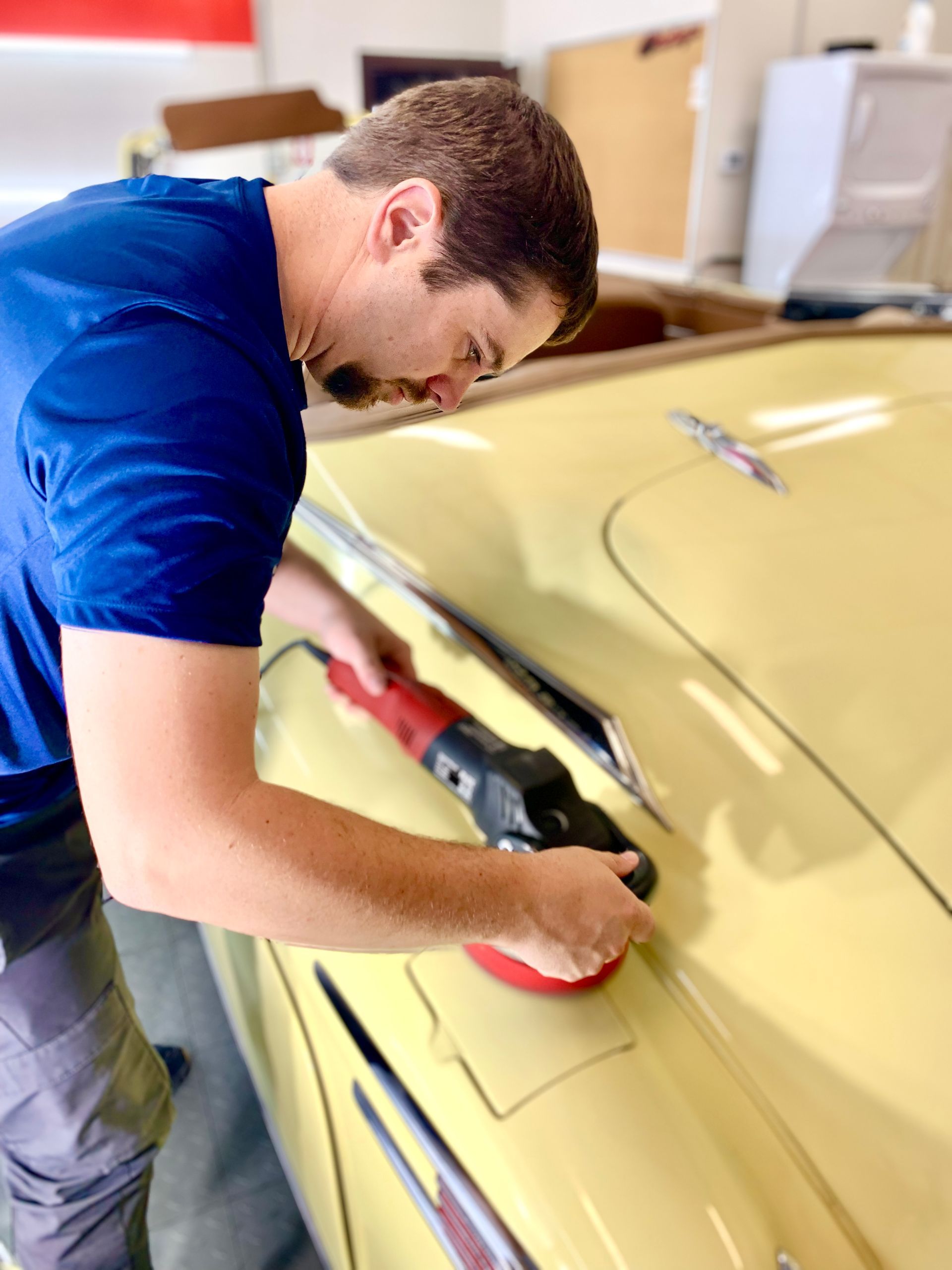 Man polishing yellow car with electric buffer in a garage.