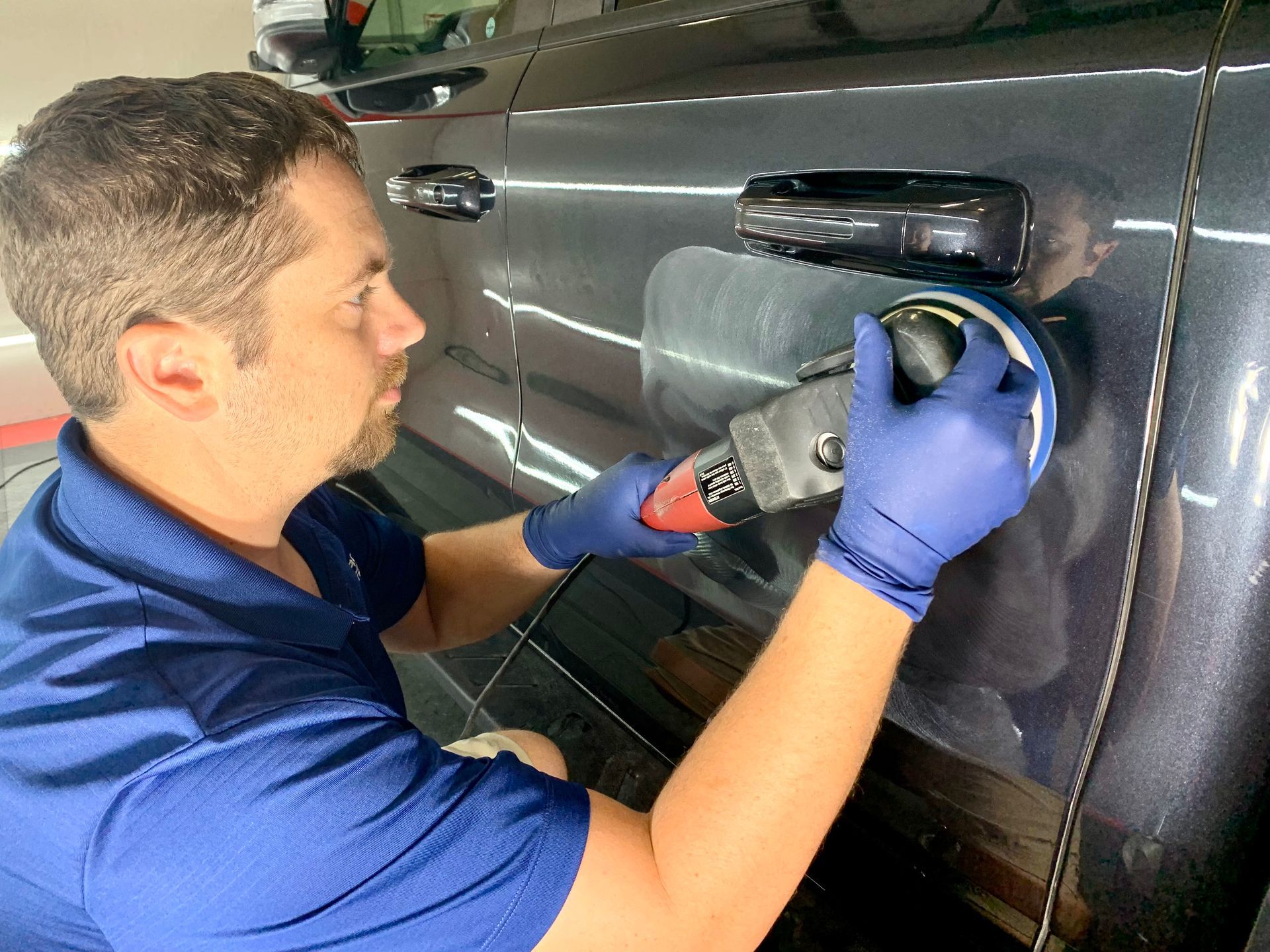 Man in blue gloves using a polisher on a gray car door, inside a garage.