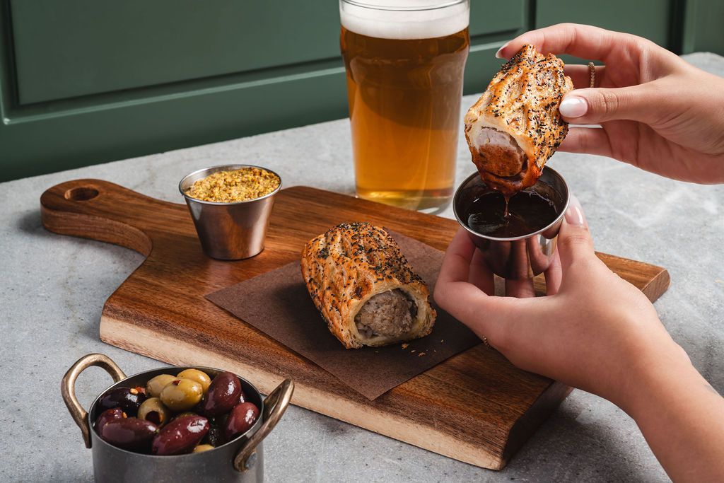 Hands dipping a sausage roll into sauvcebeside a beer and mustard on a wooden board