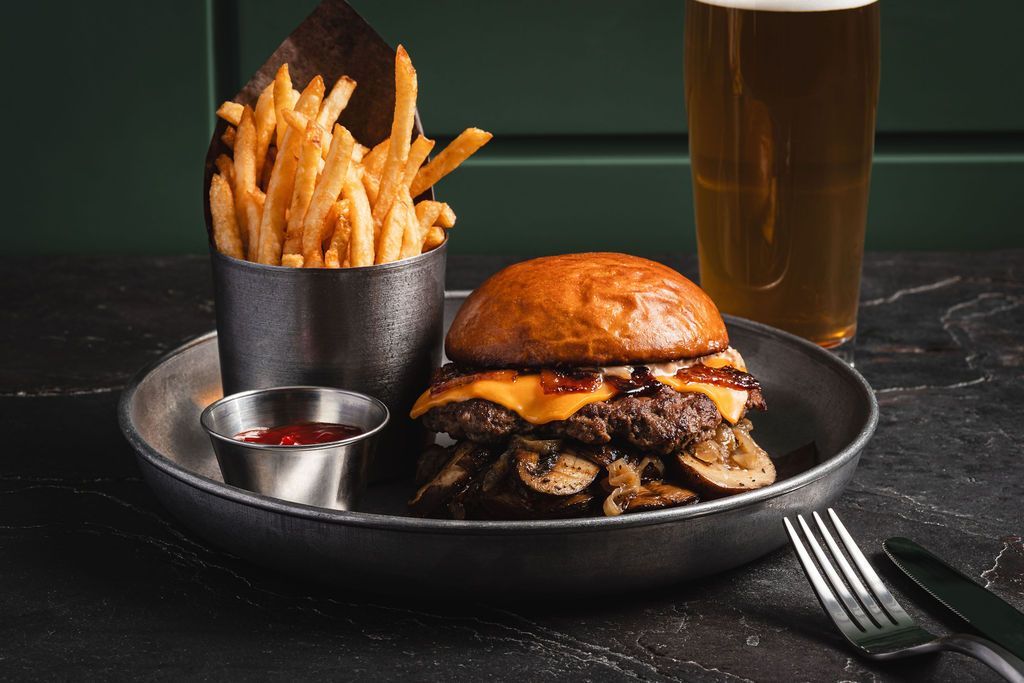 Burger with fries and a drink on a dark plate, served on a dark table with fork and knife nearby.