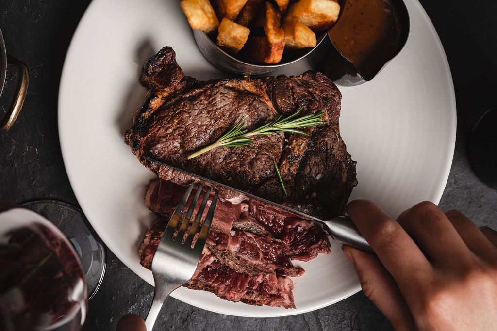 Grilled steak on a white plate with roasted potatoes and a hand cutting a slice