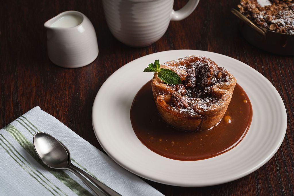 Dessert tart with sauce on a white plate beside a spoon and cream pitcher on a dark table