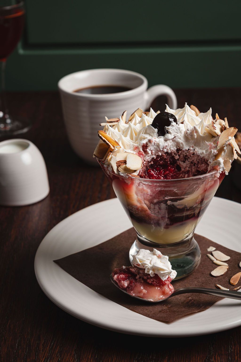 Trifle with berries and whipped cream on a white plate beside a coffee cup