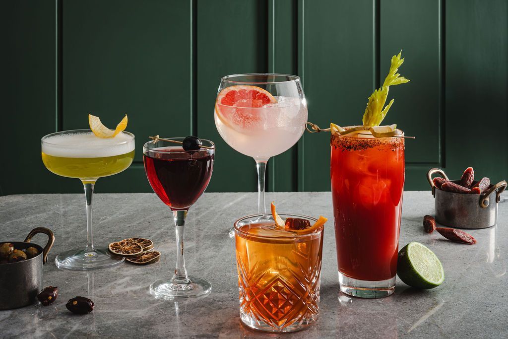 Assorted colorful cocktails arranged on a bar with citrus garnishes and a dark green backdrop