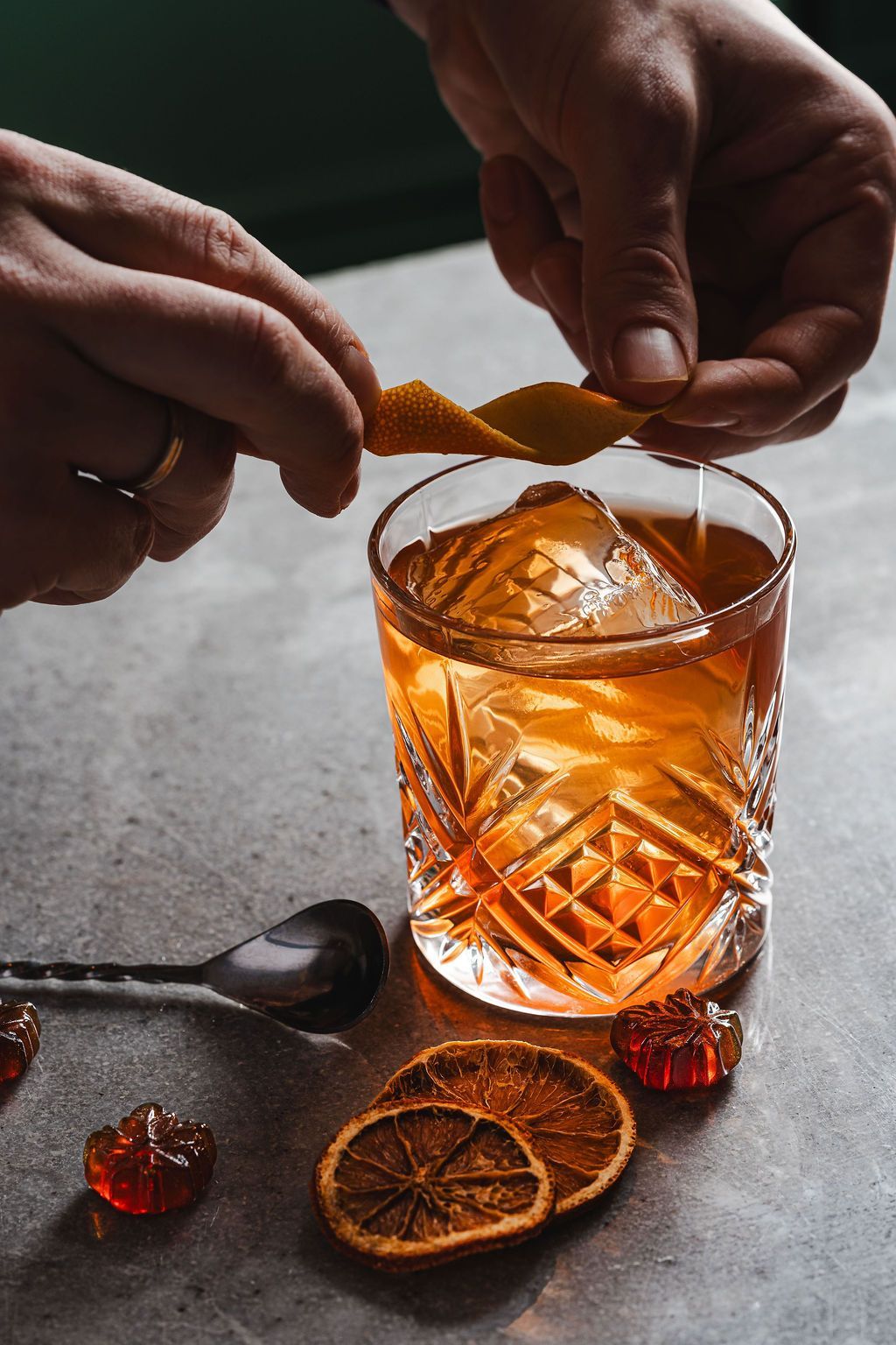 Hands garnish an amber cocktail in a cut-glass tumbler with dried orange slices on a dark table