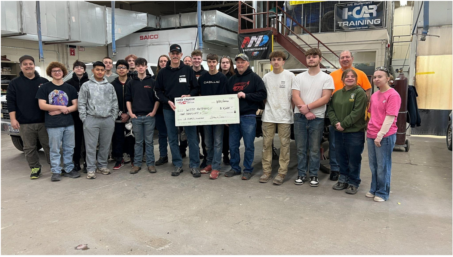 Group photo of Wayne Technical Career Center automotive students and supporters standing inside a training shop while two men hold a donation check. Automotive training signs, shop equipment, and a staircase are visible in the background.
