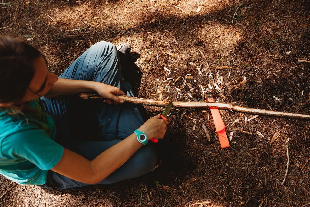A teen is sitting on the ground cutting a stick with a pair of scissors as a part of survival training in ontario