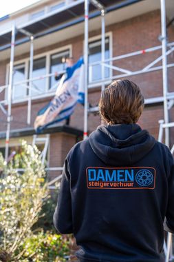 Een man in een zwarte hoodie staat voor een gebouw met een steiger.