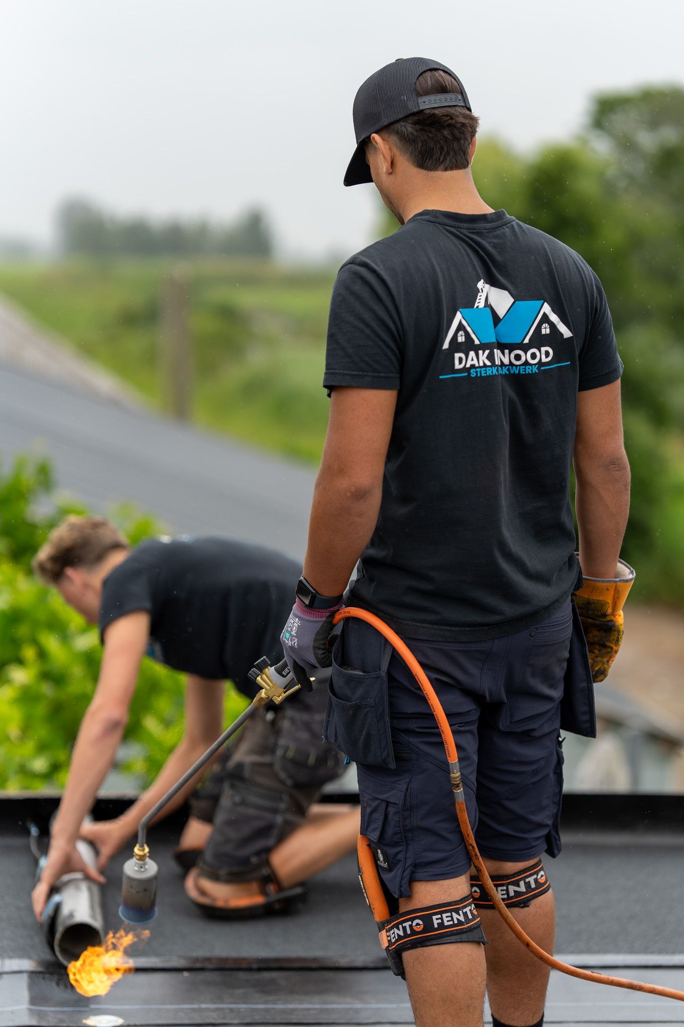 Twee mannen zijn op een dak aan het werk en een van hen houdt een slang vast.