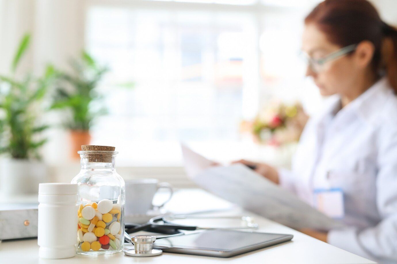 A Woman In A White Lab Coat At A Desk With Medication And A Tablet