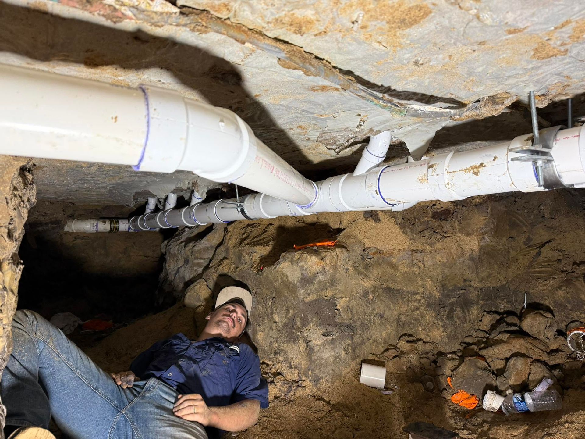 Person in a cramped crawlspace under exposed pipes, lying on the ground with tools nearby.