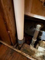 Close-up of exposed plumbing under a sink, with white and silver pipes against wood cabinetry.