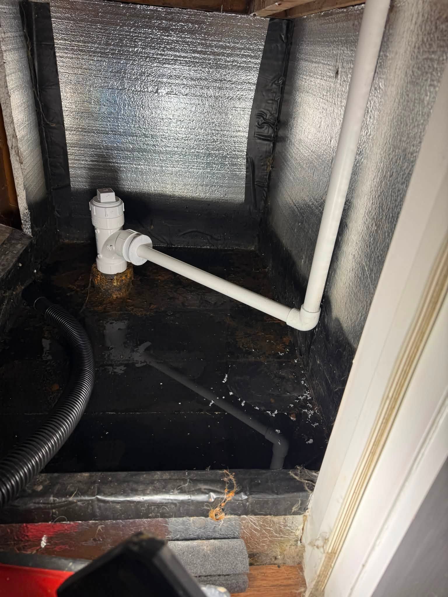 Crawlspace plumbing with white PVC pipes and a black plastic sump basin against insulated walls.
