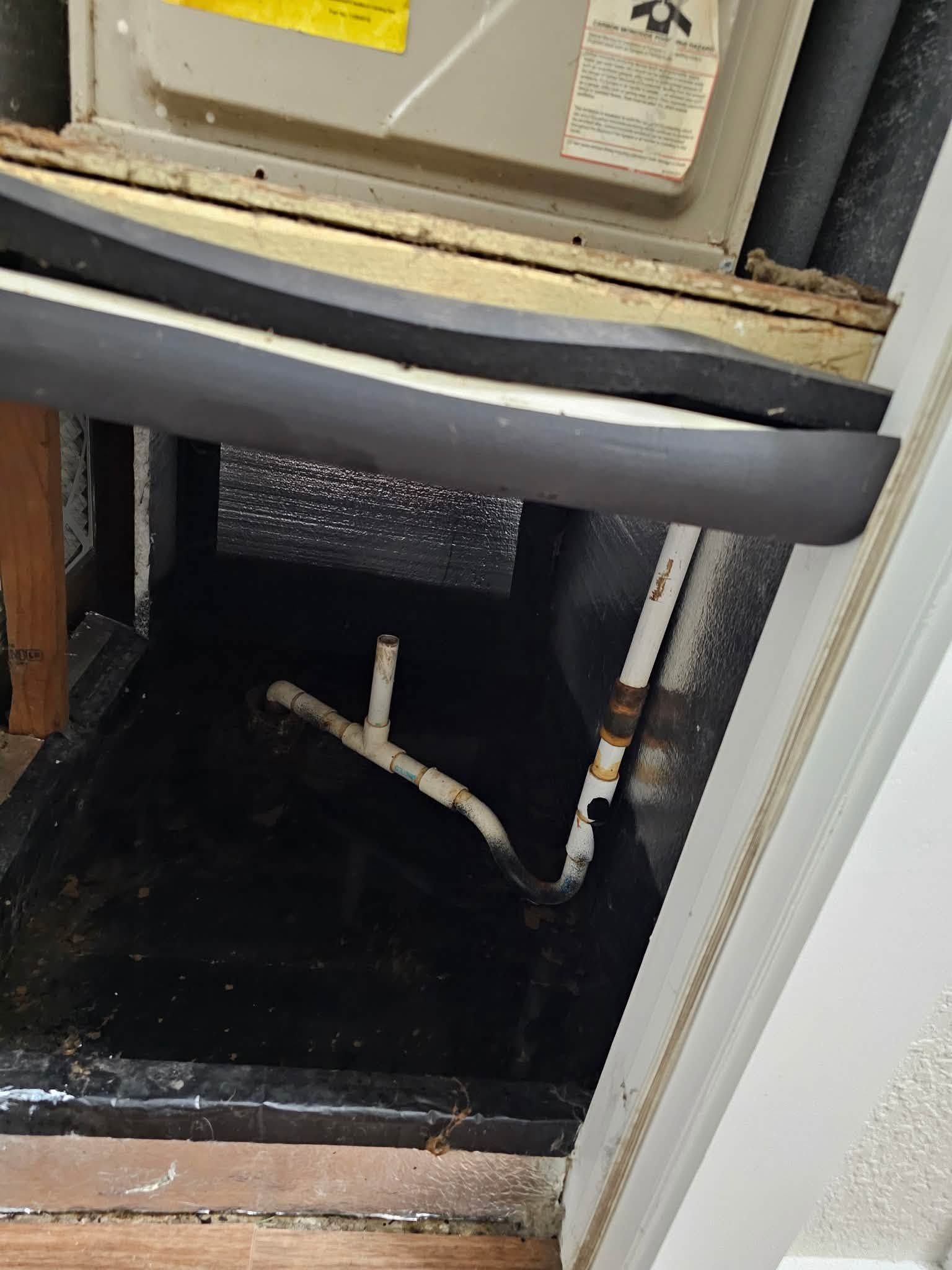 Open utility closet with HVAC unit above and exposed white drain pipes inside, framed by white trim.