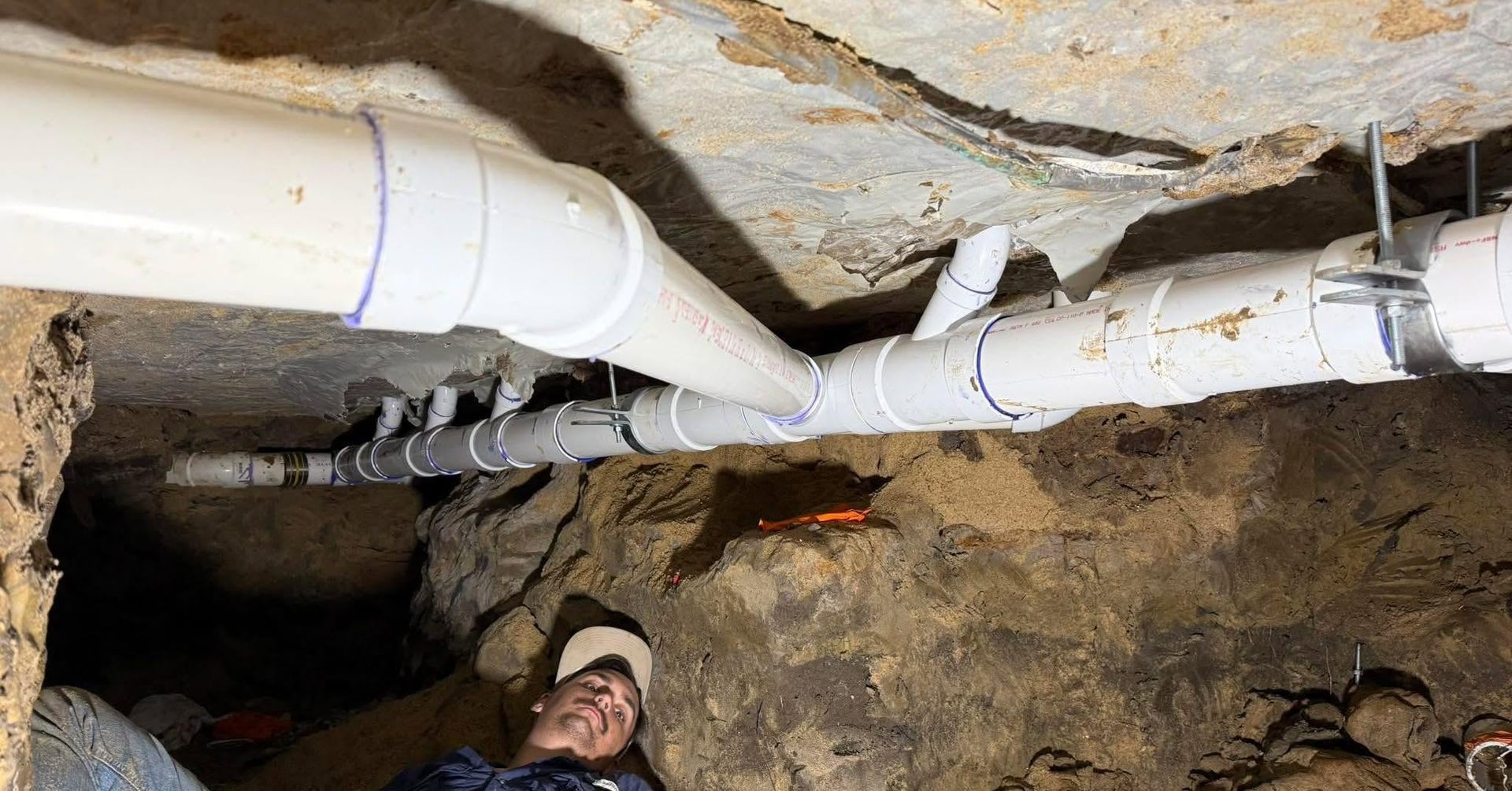 White PVC pipes run across a rocky tunnel ceiling above a worker in a cave-like space.