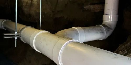 White PVC pipes and fittings in a dark crawlspace or basement, with a large horizontal drain pipe in front.
