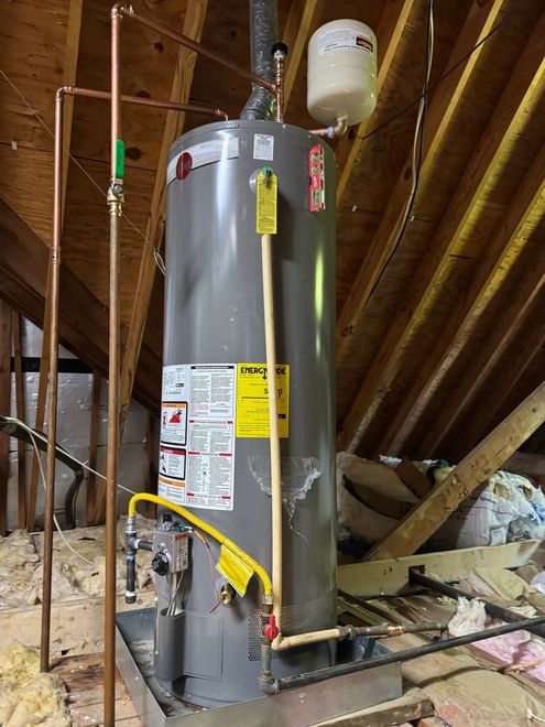 Water heater in an attic, with exposed wood rafters and plumbing pipes.