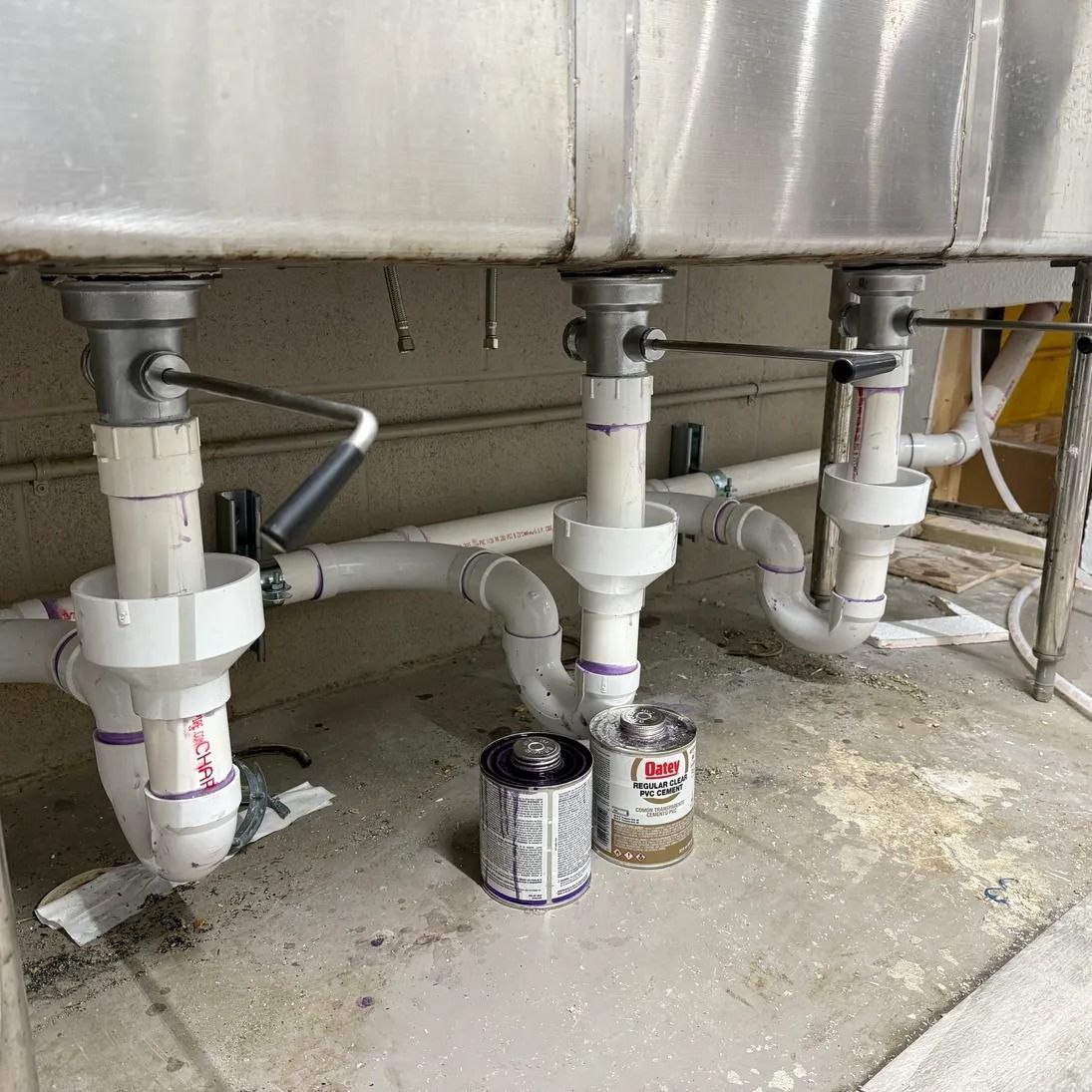 Under-sink plumbing with white PVC pipes and two small cans on a concrete floor.