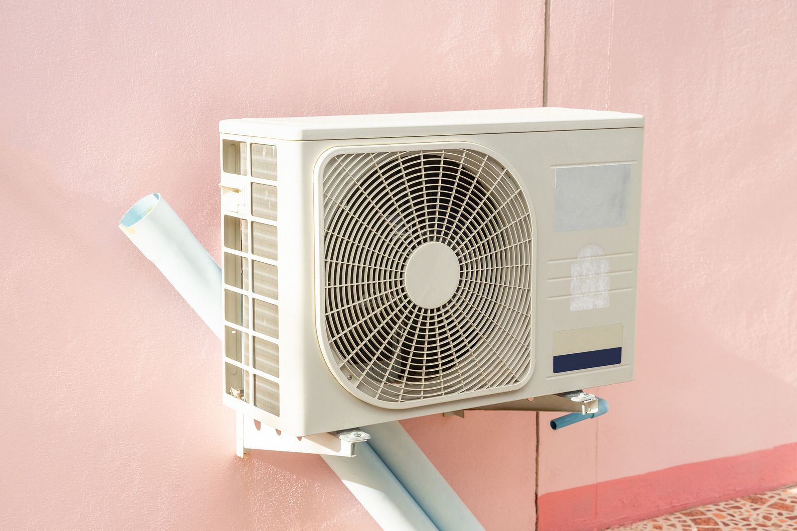 Outdoor air conditioner unit mounted on a pink wall with white piping nearby