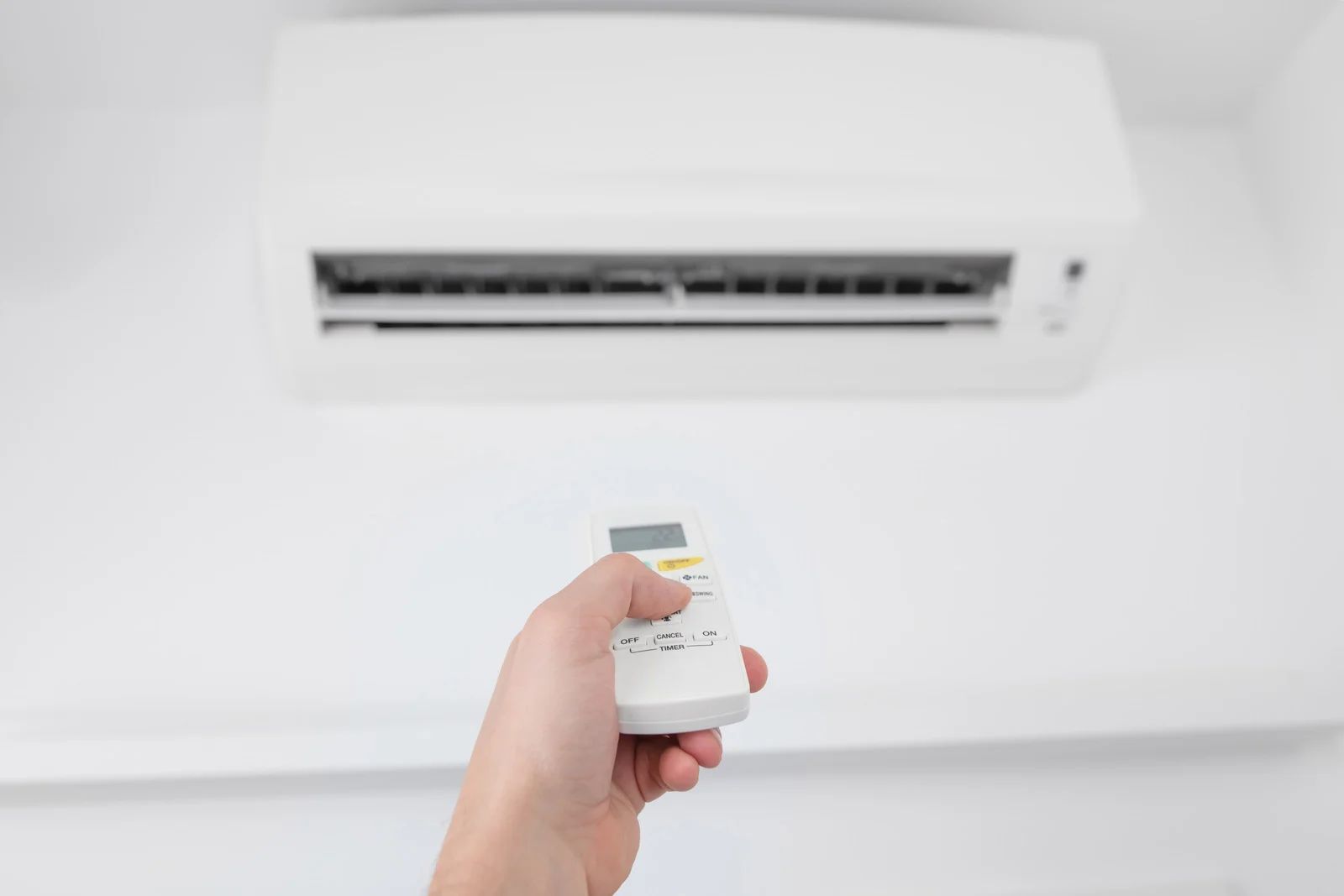 Hand holding a remote aimed at a white wall-mounted air conditioner inside a room