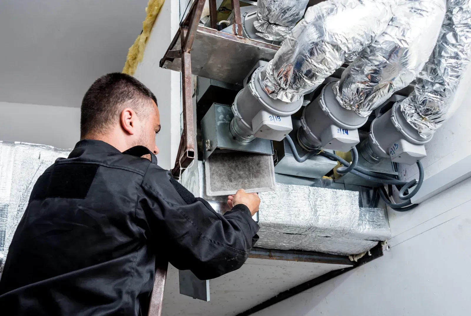 Technician inspecting ductwork in an HVAC system with insulated metal pipes and vents