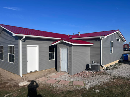 A grey, single-story building with a red metal roof, white doors, and windows, set against a clear blue sky.