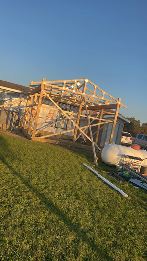 A wood-framed structure stands in a grassy yard under a clear blue sky, showing signs of damage or incomplete construction.