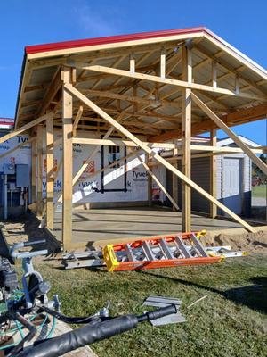 A wooden frame structure under construction with a red metal roof, a concrete slab, and a ladder on the grass in front.