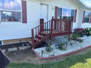 A white house with maroon shutters features a wooden porch and steps, accented by white gravel landscaping and greenery.