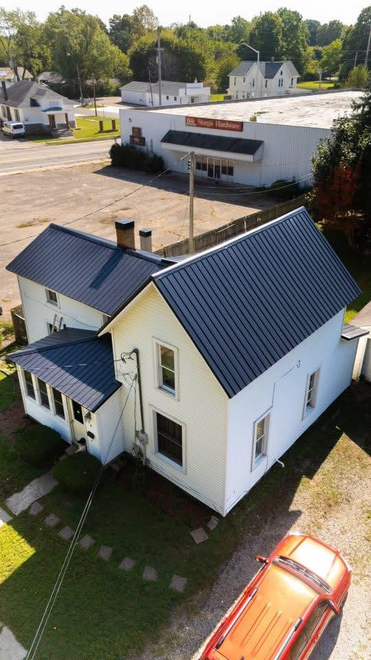Aerial view of a white, two-story house with a dark metal roof, next to a parking lot and small commercial buildings.