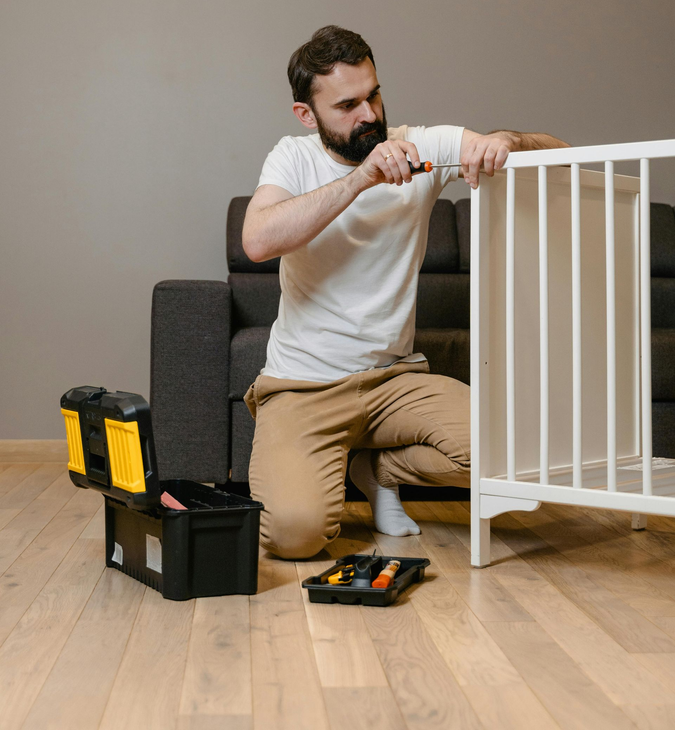 Man kneeling on a wooden floor assembling a white baby gate beside a toolbox and sofa
