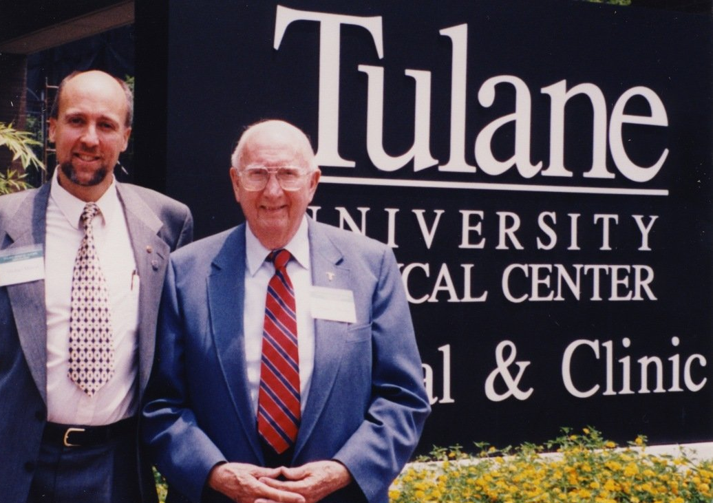 Two men in suits stand in front of a Tulane University Medical Center sign.