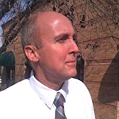 Man with a bald head wearing a white shirt and tie, looking right, with a stone structure in the background.