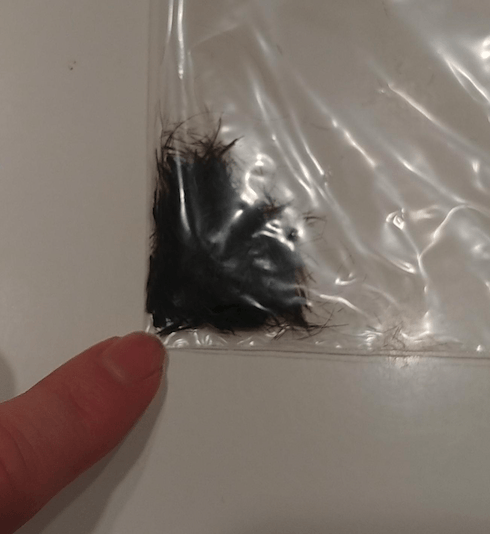Finger pointing at a small, clear plastic bag containing a clump of black hair clippings.