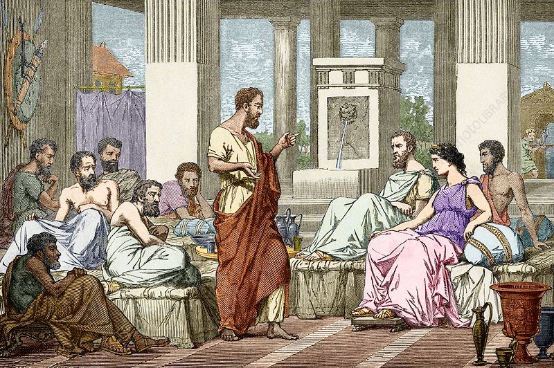 A group of people in ancient Greek attire gathered indoors listening to a man speaking, with pillars and windows.
