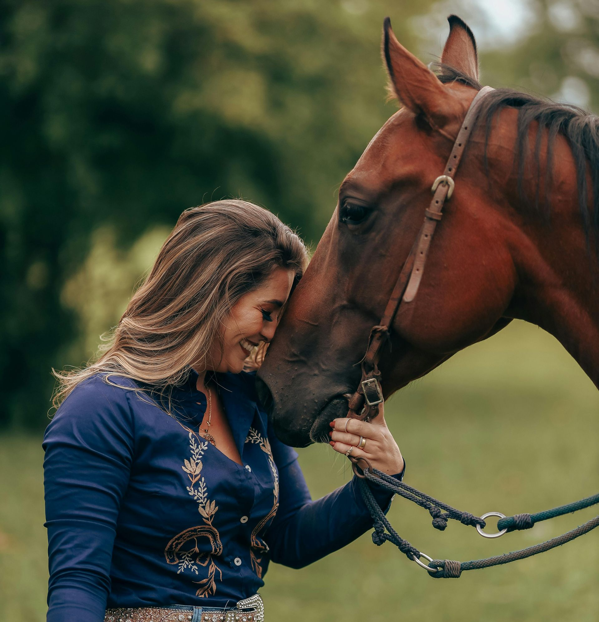 Woman in blue shirt lovingly touches a brown horse's head in a green outdoor setting.