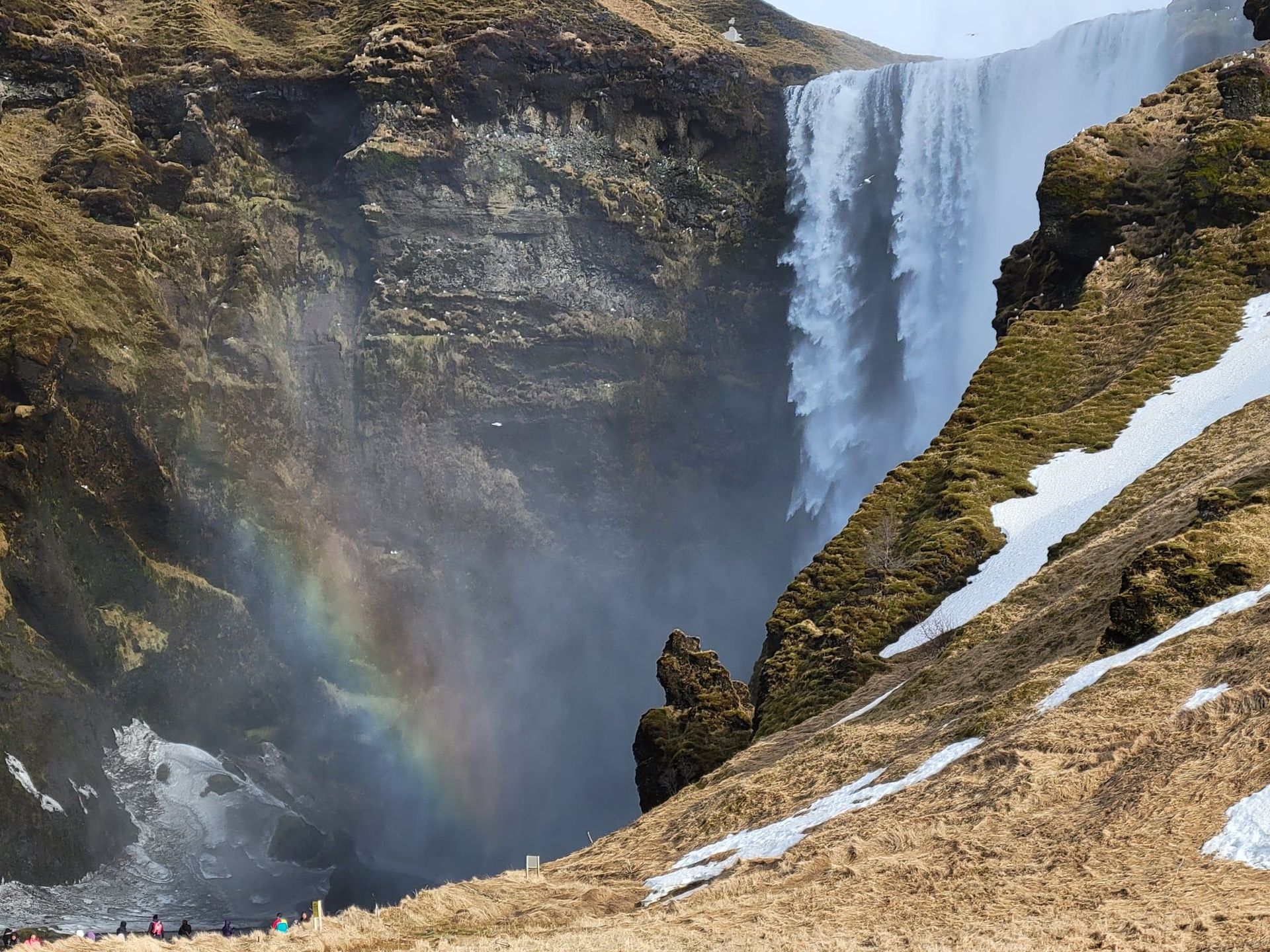 Waterfall pouring down rocky cliff with snow on the tan vegetation in the foreground. A small rainbow is visible on the left.