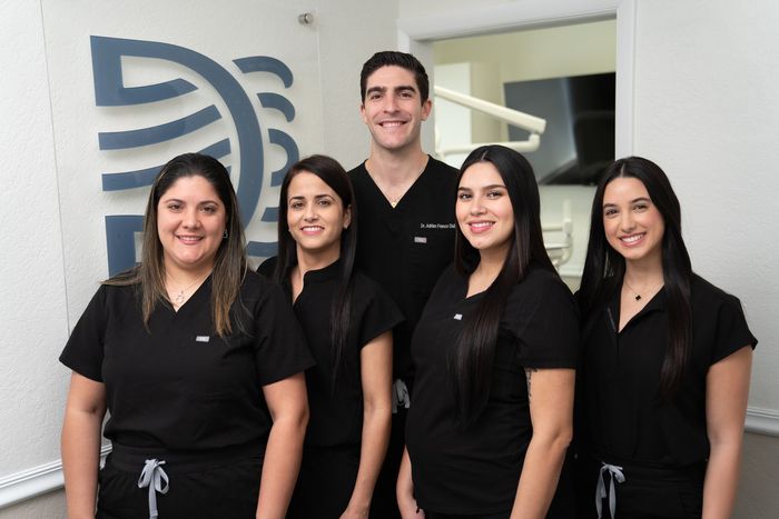 Five dental staff, wearing black scrubs, smiling in a dental office.
