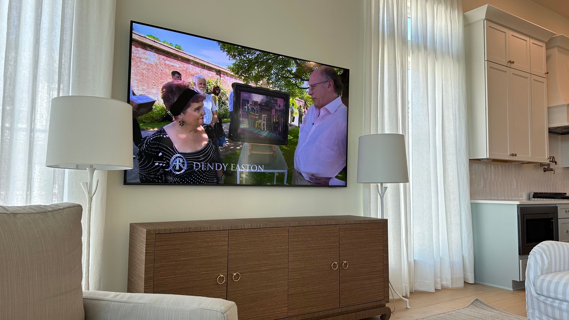 Living room with a wall-mounted TV playing a scene, flanked by floor lamps and curtains.