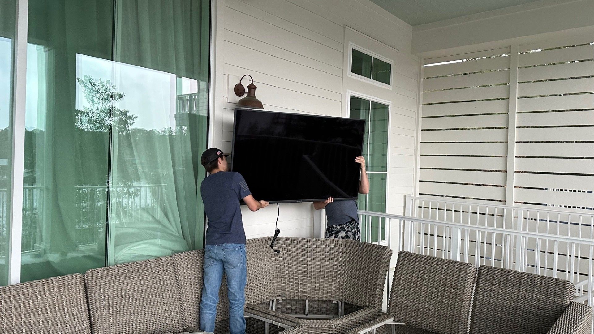 Person adjusting a large TV on a covered patio with wicker seating and green curtains