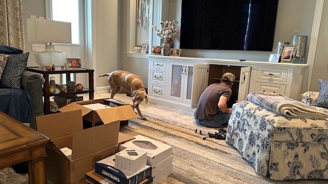 Two people unpacking boxes in a living room beside a large TV and patterned ottoman.