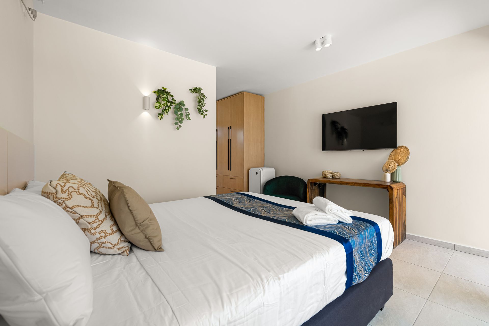 Bedroom with bed, TV, wooden desk, and wardrobe. Neutral walls, white bedding, and green plant decorations.