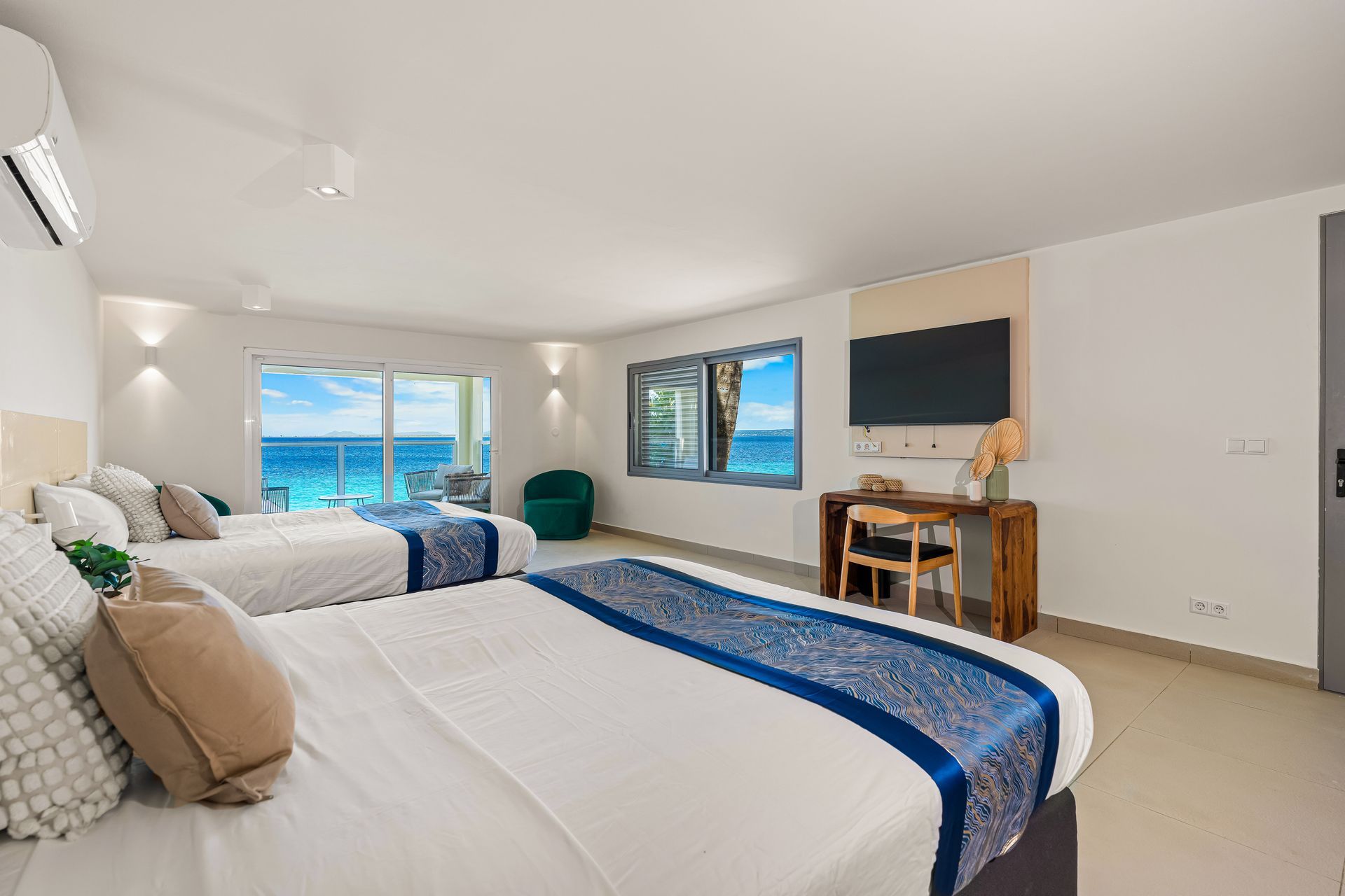 Bright hotel room with two beds, ocean view, desk, and wall-mounted TV