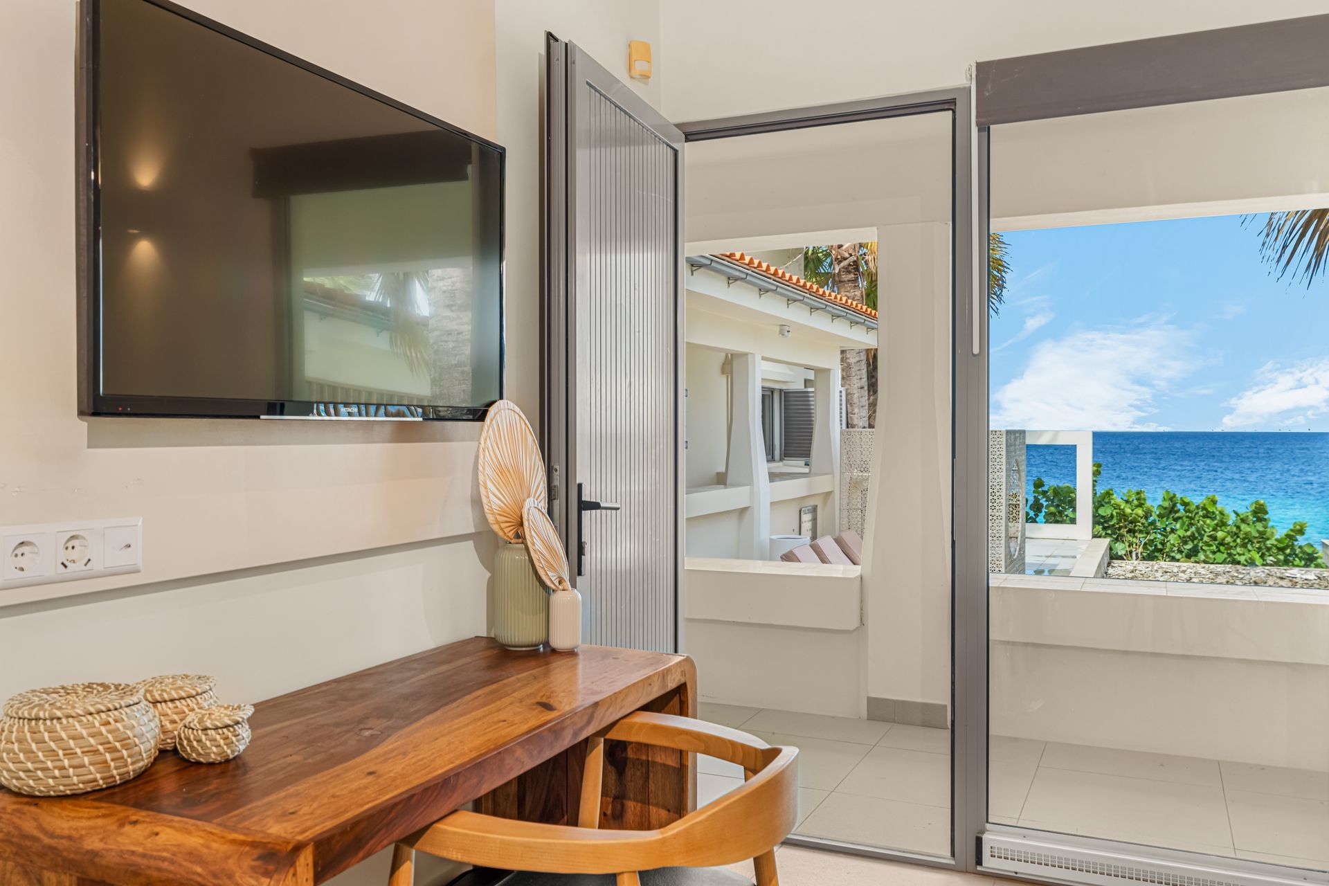 A room with a desk, TV, and balcony overlooking the ocean.