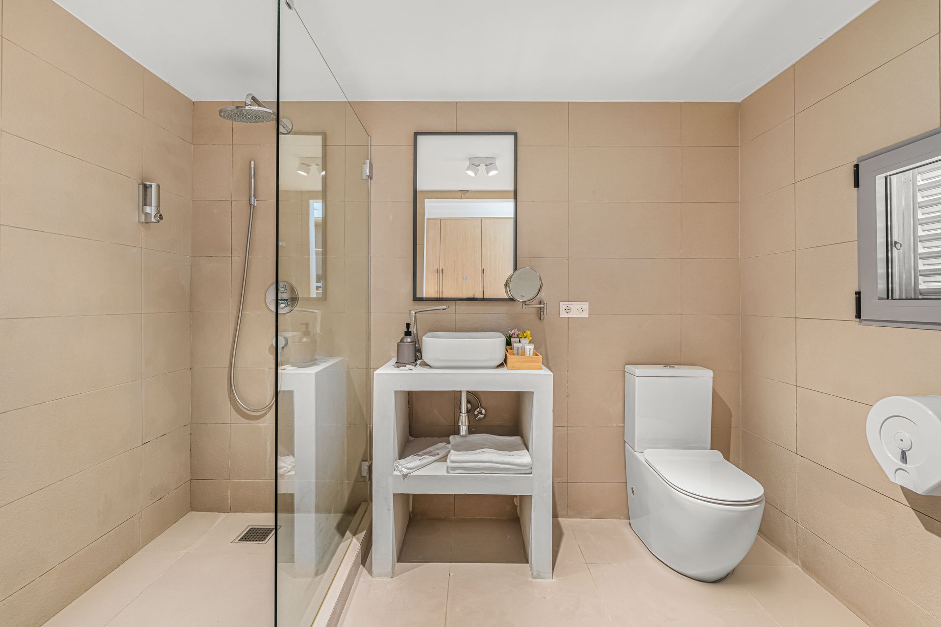 Beige-tiled bathroom with a glass shower, white sink, toilet, and a small window; a minimalist aesthetic.